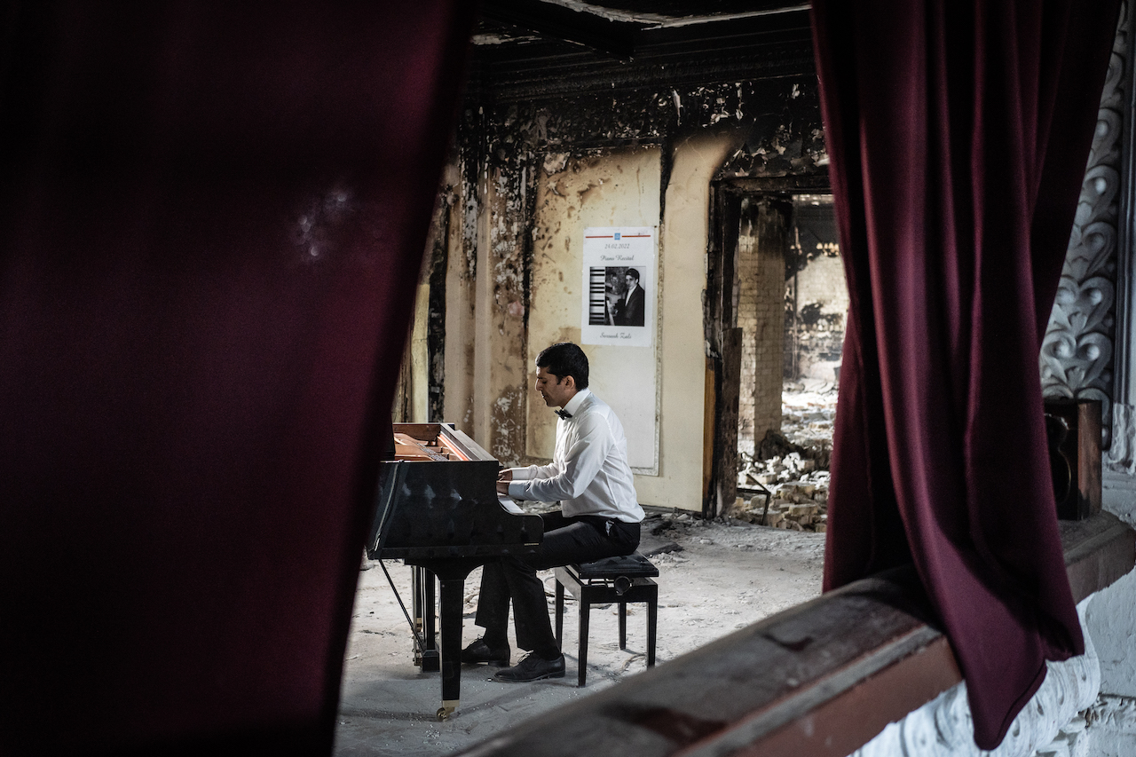 A man in a white shirt sits at a piano in a bombed-out room