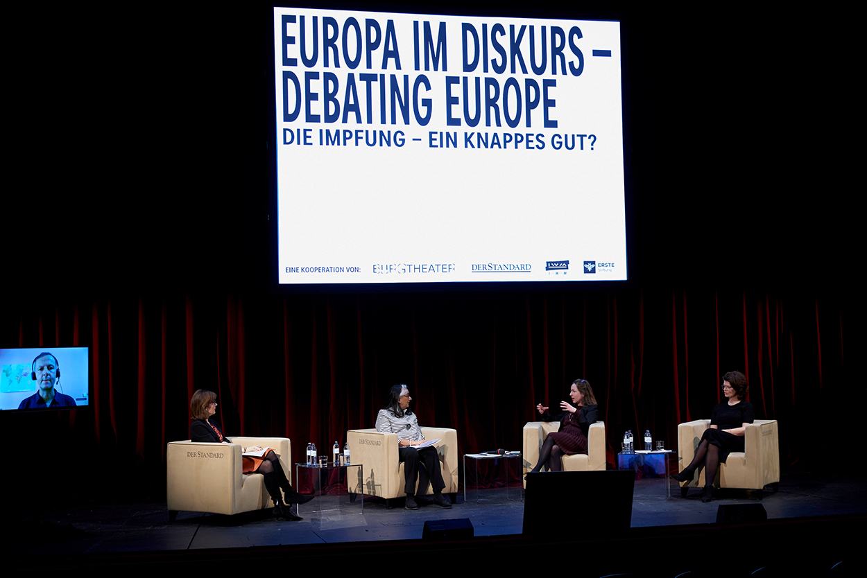 The stage of the Burgtheater, with the panelists seated at a distance from each other. A large screen behind bears the name of the event in black and white.