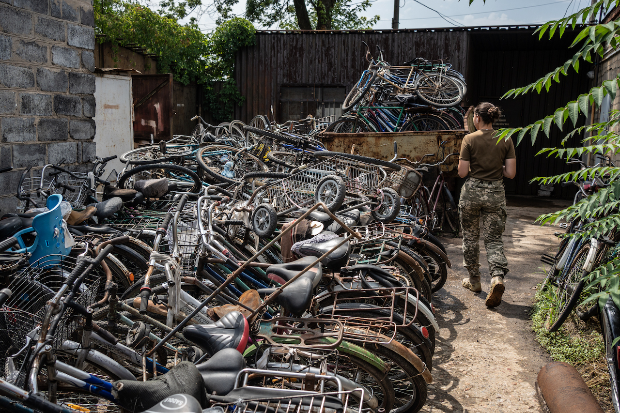Вozens of broken bicycles piled in a heap, a Ukrainian female soldier passes by