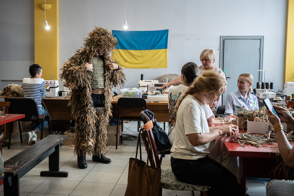 A man tries on camouflage, women are around with sewing machines