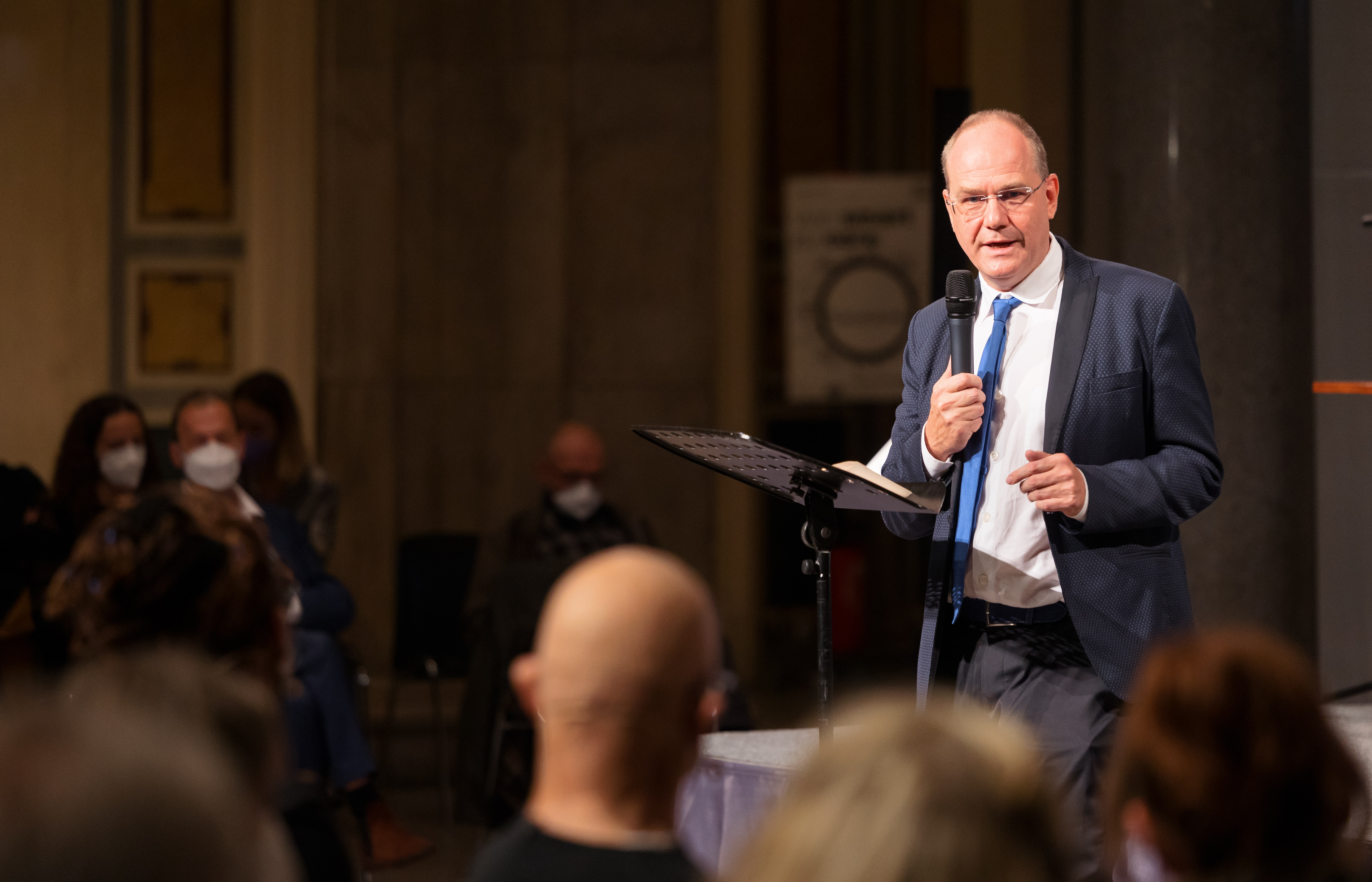 Ludger Hagedorn speaks at the MAC in front of a crowd, wearing a suit and blue tie