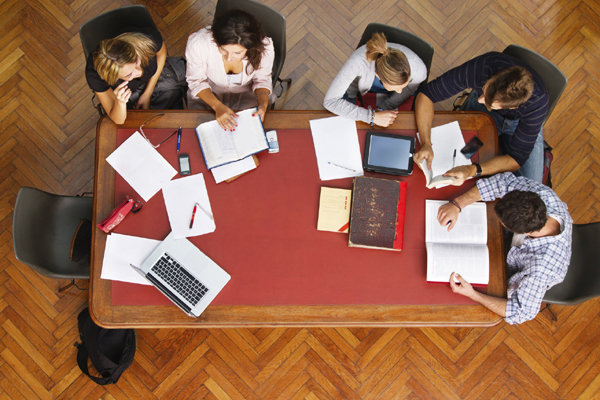 An aerial view of a red table, fellows are sitting around it with notebooks and books on top of the table, deep in discussion.