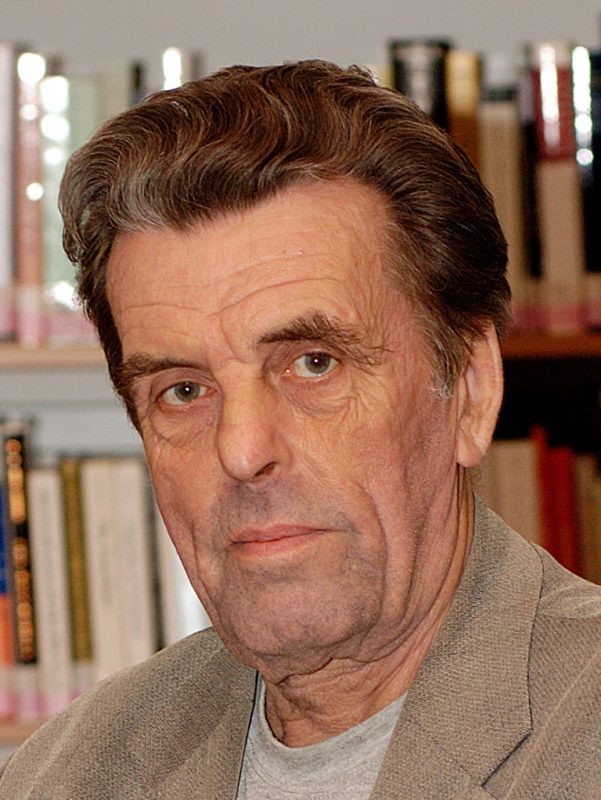 Headshot of Jan Sokol, in front of a book case wearing a light coloured jacket