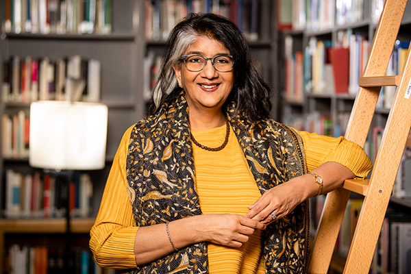 Shalini Randeria wearing an yellow top and scarf, leans on a ladder in the IWM library