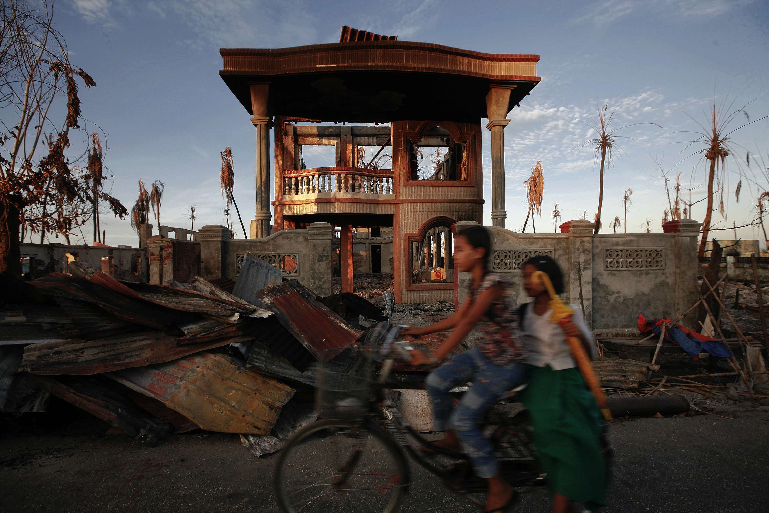 Girls ride a moped past a burnt house during the violence at East Pikesake ward, in Kyaukphyu November 6, 2012. Photo: STRINGER / REUTERS / picturedesk.com