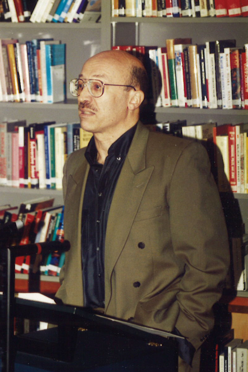 Tony Judt standing at a podium in the IWM library in front of shelves of books giving a lecture. He is wearing a dark shirt and brown suit jacket.