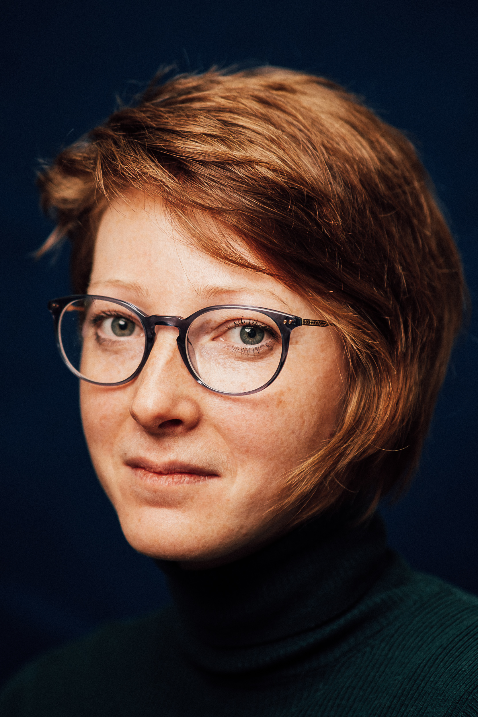 Katerina Koci with a black background, wearing a black turtleneck and glasses looking into the camera