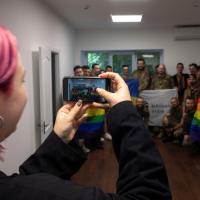 A woman with pink hair taking a picture of a group of military personnel holding LGBTQ+ flags