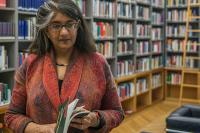 Shalini Randeria, wearing a red jacket, is standing in the IWM library holding a book.