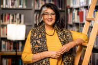 Shalini Randeria in a yellow top leans on the library ladder in the IWM library, surrounded by books