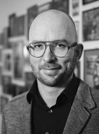 headshot of Felix Ackermann in black and white. He is wearing a dark shirt and light jacket standing in front of a wall of framed pictures.