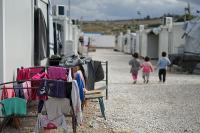 A displacement camp comprising 2 rows of container housing and a temporary road between them. in the foreground is some brightly coloured washing. in the background three small children walk away from the camera.