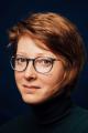 Katerina Koci with a black background, wearing a black turtleneck and glasses looking into the camera