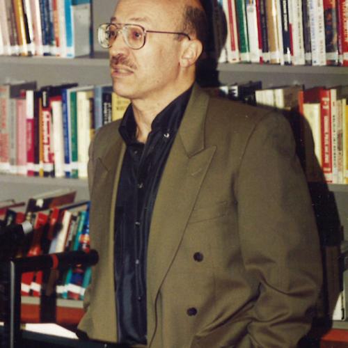 Tony Judt standing at a podium in the IWM library in front of shelves of books giving a lecture. He is wearing a dark shirt and brown suit jacket.