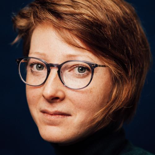 Katerina Koci with a black background, wearing a black turtleneck and glasses looking into the camera